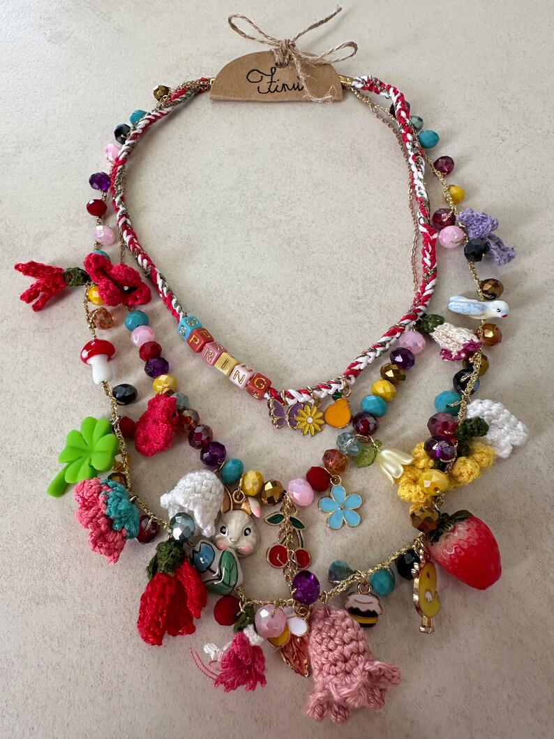 May include: Multi-layered necklace with a spring theme. It features crocheted flowers, a bunny charm, a strawberry charm, and beads in various colors. The necklace also includes a tag that says "Spring".