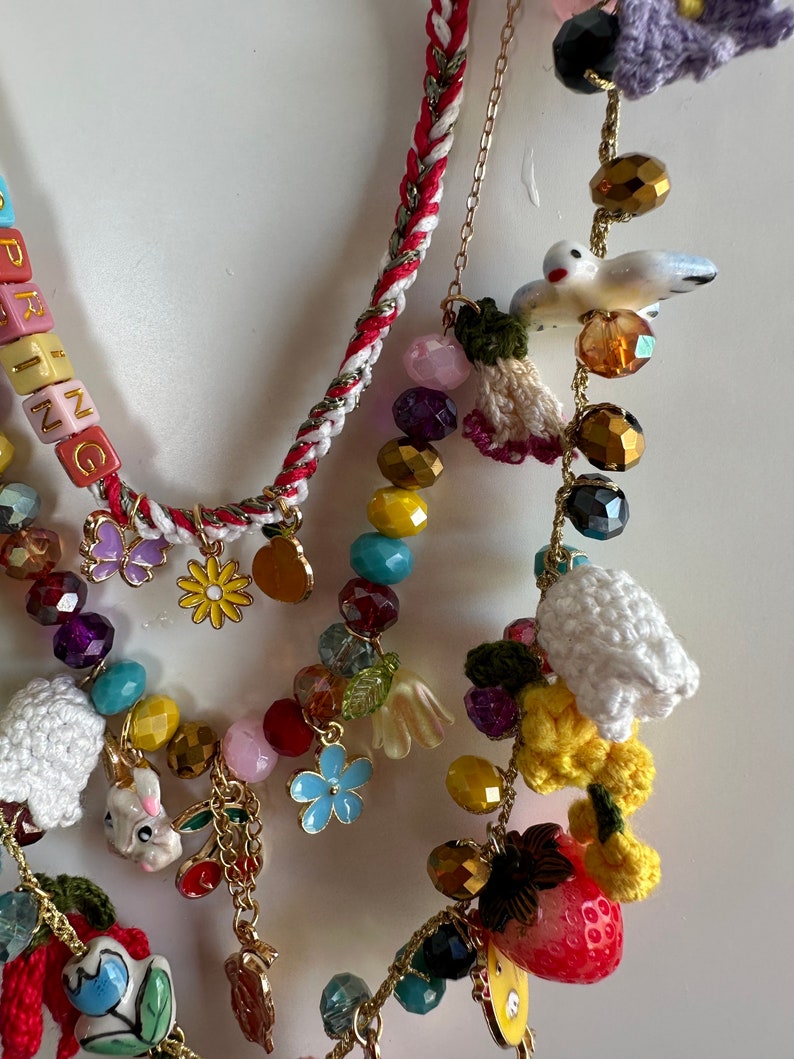 May include: A multi-strand necklace with colorful beads, charms, and crocheted elements. The necklace features a variety of charms, including flowers, birds, strawberries, and a bunny. The necklace also includes a beaded section with the word "SPRING" spelled out in colorful letters.