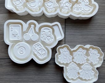 May include: Three white plastic cookie cutters featuring various designs. One cutter has five ducks in winter attire, another features casino-themed icons like a playing card, cherries, a slot machine, an eight ball, and dice, and the third has five ghosts.