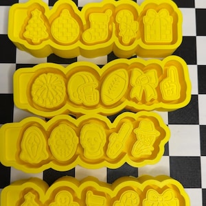 Puede incluir: Cuatro tiras de cortadores de galletas de plástico amarillo, cada una con cinco formas distintas. Los diseños incluyen iconos navideños como árboles, adornos, calcetines, bastones de caramelo, regalos, motivos deportivos como balones y cascos, y máscaras de terror.