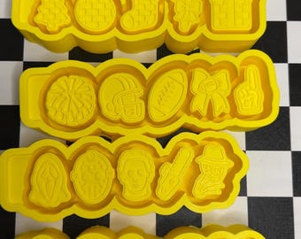 May include: Four yellow plastic cookie cutter strips, each containing five distinct shapes. The designs include holiday icons like trees, ornaments, stockings, candy canes, gifts, sports motifs like footballs and helmets, and various horror movie character masks.