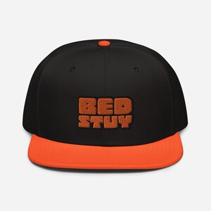 Puede incluir: Una gorra de béisbol negra y naranja con el texto "BED-STUY" bordado en naranja en la parte delantera.