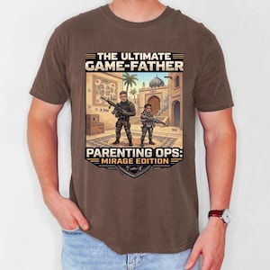 May include: Brown t-shirt featuring a graphic with the text "THE ULTIMATE GAME-FATHER" and "PARENTING OPS: MIRAGE EDITION". The graphic depicts a father and child holding weapons in a desert scene. The shirt is worn by a person.