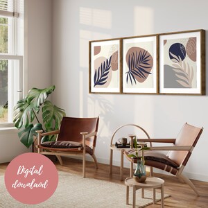 May include: Three framed prints with abstract designs featuring leaves and organic shapes in shades of beige, blue, and grey. The prints are hung on a white wall in a living room setting with two leather chairs and a wooden coffee table.