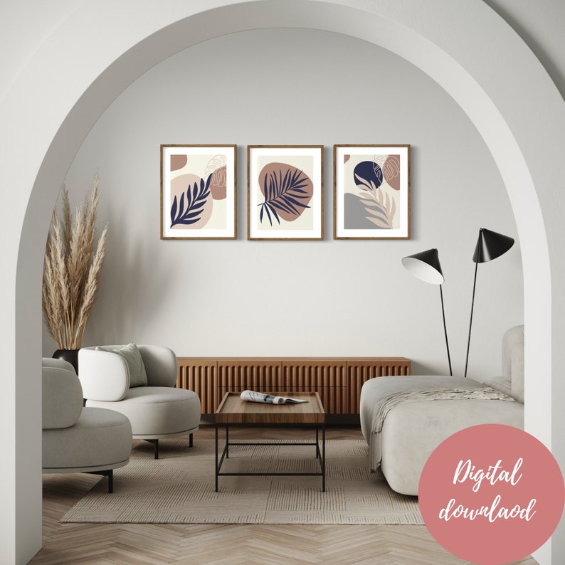 May include: Three framed prints with abstract designs featuring blue and brown leaves on a cream background. The prints are hung on a white wall in a modern living room setting.