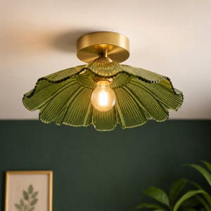 Peut inclure: Un plafonnier en verre vert avec un abat-jour à volants en forme de pétale. Le luminaire a une base de couleur laiton et une ampoule visible, de couleur chaude. Le luminaire est monté sur un plafond blanc.