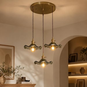 Puede incluir: Una lámpara colgante de tres luces con acabado de latón y tres pantallas de cristal gris. Cada pantalla tiene un borde con volantes y una bombilla de estilo vintage. La lámpara cuelga de una placa de techo de latón con cables trenzados marrones.