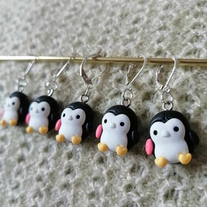 Op de afbeelding: Vijf paar hangende oorbellen met zwarte en witte pinguïnbedels met roze en gele accenten. De pinguïns zitten met hun voeten bij elkaar en hun vleugels uitgespreid.
