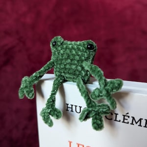 Puede incluir: Un marcapáginas de rana verde tejido a crochet, sentado encima de un libro. El libro tiene el título "Les Lapin ne mangent de...