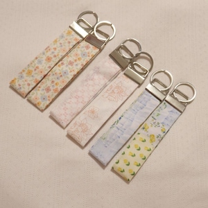May include: A collection of fabric keychains with floral patterns in pastel colors. Each keychain has a silver-colored metal clasp and ring. The patterns include small flowers in pink, yellow, and blue, and a light pink floral design.