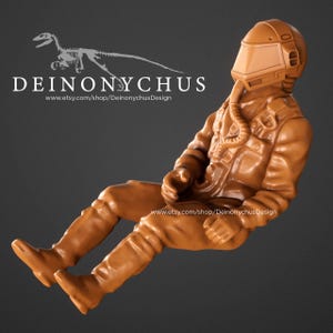 May include: A brown figurine of a seated pilot wearing a helmet and flight suit. The figure is in a seated position with legs extended. The text "DEINONYCHUS" and a website address are visible.