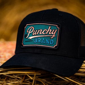Peut inclure: Casquette de camionneur noire avec un écusson rectangulaire. L'écusson affiche le mot "Punchy" en écriture cursive, au-dessus du mot "BRAND". L'écusson a un fond vert d'eau avec des accents roses et blancs.
