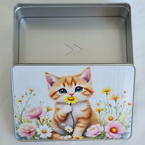 Könnte beinhalten: Eine rechteckige Metallbox mit einem Klappdeckel. Der Deckel zeigt eine farbenfrohe Illustration eines orangefarbenen Tabby-Kätzchens, das eine gelbe Blume hält, umgeben von rosa und weißen Blumen und grünem Laub.