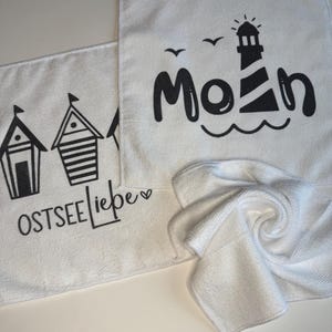 May include: White microfiber cloths with black graphic designs. One cloth features beach huts and the text "OSTSEE Liebe". Another cloth displays a lighthouse, waves, and the word "Moin". A third cloth is folded.