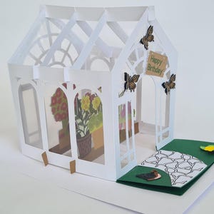 Puede incluir: Tarjeta desplegable de papel blanco con forma de invernadero, con base verde. La tarjeta presenta un diseño detallado con mariposas y la frase "Happy Birthday". En el interior, imágenes de flores en macetas.