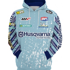 Op de afbeelding: Een lichtblauwe hoodie met een wit en blauw gestreept patroon op de mouwen. De hoodie heeft verschillende logo's die op de voorkant zijn gedrukt, waaronder "Husqvarna Factory Racing", "Michelin", "100%", "ProTaper", "Motorex" en "Rockstar".