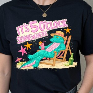 Könnte beinhalten: Schwarzes T-Shirt mit einem Cartoon-Alligator, der sich in einem Liegestuhl entspannt und einen Cocktail hält. Das Design enthält den Text "IT'S 5 O'CLOCK SOMEWHERE" in Rosa und Weiß, mit Strandmotiven.