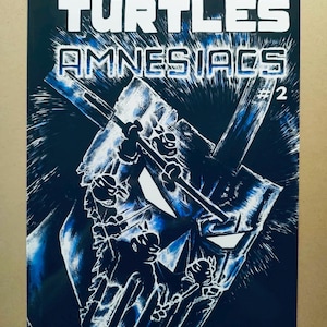 May include: Comic book cover for "Teenage Mutant Ninja Turtles Amnesiacs #2". The artwork features a dark blue background with white and blue illustrations of the turtles and a large, stylized mask. The title and creators' names are at the top.