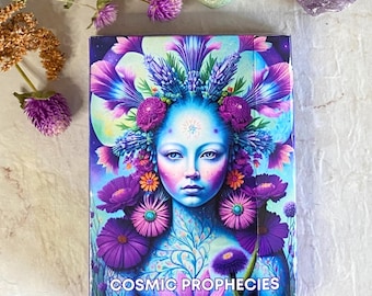 May include: A box of Cosmic Prophecies Oracle Cards featuring a colorful illustration of a woman with a floral crown and a blue face. The box is decorated with purple, blue, and green flowers.