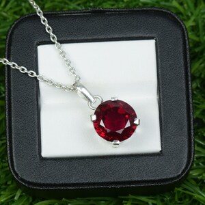 May include: A silver pendant necklace featuring a large, round, deep red gemstone. The gemstone is set in a silver bezel and hangs from a delicate silver chain. The necklace is displayed in a black jewelry box with a white interior.
