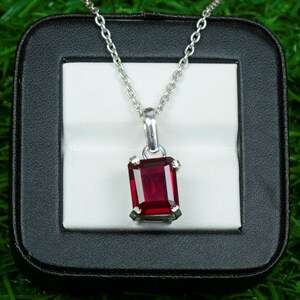Peut inclure: Un collier en argent avec un pendentif rectangulaire en pierre précieuse rouge foncé. Le pendentif est serti dans un cadre argenté et suspendu à une fine chaîne en argent. Le collier est présenté dans un écrin noir avec un intérieur blanc, sur fond vert.