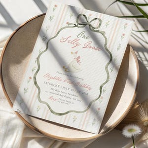 May include: A whimsical invitation for a first birthday celebration. The design features a watercolor illustration of a goose wearing a party hat, with the text "One Silly Goose." The invitation is set on a striped background with floral accents.