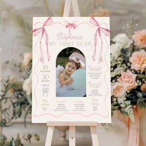 May include: A personalized first birthday sign on an easel. The sign features a photo of a baby, with stats including 18 pounds and 30 inches. The sign is decorated with pink bows and ribbons, and includes the text "Stephanie MY FIRST YEAR."