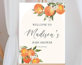 May include: A baby shower welcome sign on an easel. The sign features watercolor illustrations of oranges with green leaves and white blossoms. The text reads "Welcome to Madison's Baby Shower April 20th 2024."