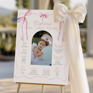 May include: A personalized first birthday sign with a photo of a baby girl. The sign includes the baby's name, weight (18 pounds), height (30 inches), and favorite things. Pink bows and ribbons decorate the sign.