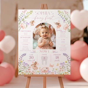 May include: A personalized sign on an easel, featuring a photo of a baby girl. The sign includes the text "Sophia's Fairy First Year," along with details like height (30 inches), weight (18 pounds), and favorite treats. Floral and fairy illustrations decorate the sign.