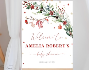 Può includere: Un cartello di benvenuto bianco con una ghirlanda acquerellata rossa e verde, fiocchi di neve e una corda di vestiti per bambini appesa a un ramo. Il testo recita "Benvenuti al Baby Shower di Amelia Robert il 20 dicembre".