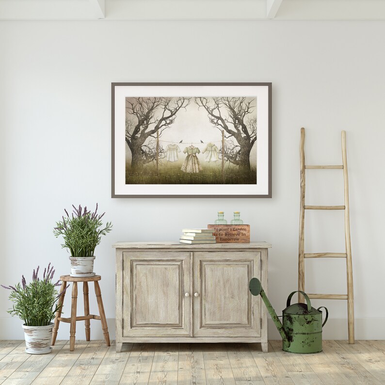 May include: A framed print of a vintage illustration depicting two dresses hanging on a clothesline in a forest setting. The print is displayed on a white wall above a wooden cabinet with two doors. There are two potted plants and a watering can on the floor.