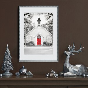 K&ouml;nnte beinhalten: Ein gerahmtes Schwarzwei&szlig;foto einer wei&szlig;en Kirche mit einer roten T&uuml;r, umgeben von schneebedeckten B&auml;umen. Das Foto ist auf einem braunen Kaminsims mit silbernen Weihnachtsdekorationen ausgestellt, darunter ein kleiner silberner Weihnachtsbaum, ein silberner Christbaumschmuck und ein silbernes Rentier.