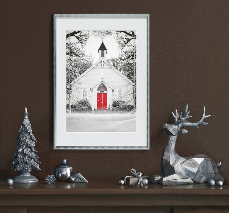 K&ouml;nnte beinhalten: Ein gerahmtes Schwarzwei&szlig;foto einer wei&szlig;en Kirche mit einer roten T&uuml;r, umgeben von schneebedeckten B&auml;umen. Das Foto ist auf einem braunen Kaminsims mit silbernen Weihnachtsdekorationen ausgestellt, darunter ein kleiner silberner Weihnachtsbaum, ein silberner Christbaumschmuck und ein silbernes Rentier.