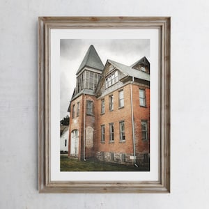 May include: A framed print of a historic brick building with a distinctive tower and multiple windows. The building has a weathered appearance, with a gray roof and a white building in the background. The image is set in a rustic wooden frame.