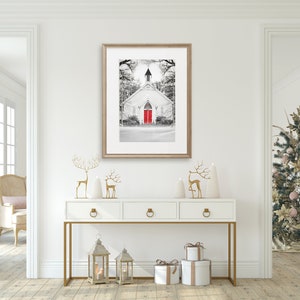 K&ouml;nnte beinhalten: Eine wei&szlig;e Kirche im Farmhouse-Stil mit einer leuchtend roten T&uuml;r, umgeben von Schnee und B&auml;umen, in einem Schwarzwei&szlig;foto. Das Foto ist gerahmt und h&auml;ngt an einer wei&szlig;en Wand &uuml;ber einem wei&szlig;en Konsolentisch mit goldenen Akzenten. Der Konsolentisch ist mit goldenen Rentierfiguren, wei&szlig;en Weihnachtsb&auml;umen und goldenen Laternen dekoriert.