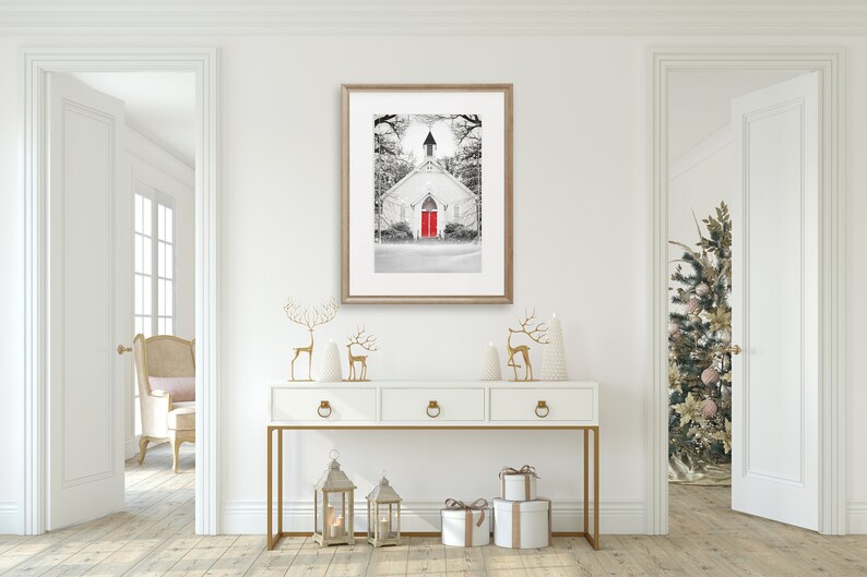 K&ouml;nnte beinhalten: Eine wei&szlig;e Kirche im Farmhouse-Stil mit einer leuchtend roten T&uuml;r, umgeben von Schnee und B&auml;umen, in einem Schwarzwei&szlig;foto. Das Foto ist gerahmt und h&auml;ngt an einer wei&szlig;en Wand &uuml;ber einem wei&szlig;en Konsolentisch mit goldenen Akzenten. Der Konsolentisch ist mit goldenen Rentierfiguren, wei&szlig;en Weihnachtsb&auml;umen und goldenen Laternen dekoriert.