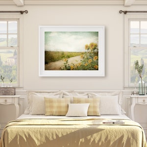 Puede incluir: Una impresi&oacute;n enmarcada en blanco de una fotograf&iacute;a de estilo vintage de un camino de tierra que conduce a trav&eacute;s de un campo de girasoles. El cielo es azul claro con nubes blancas. Los girasoles est&aacute;n en flor y el campo es verde.
