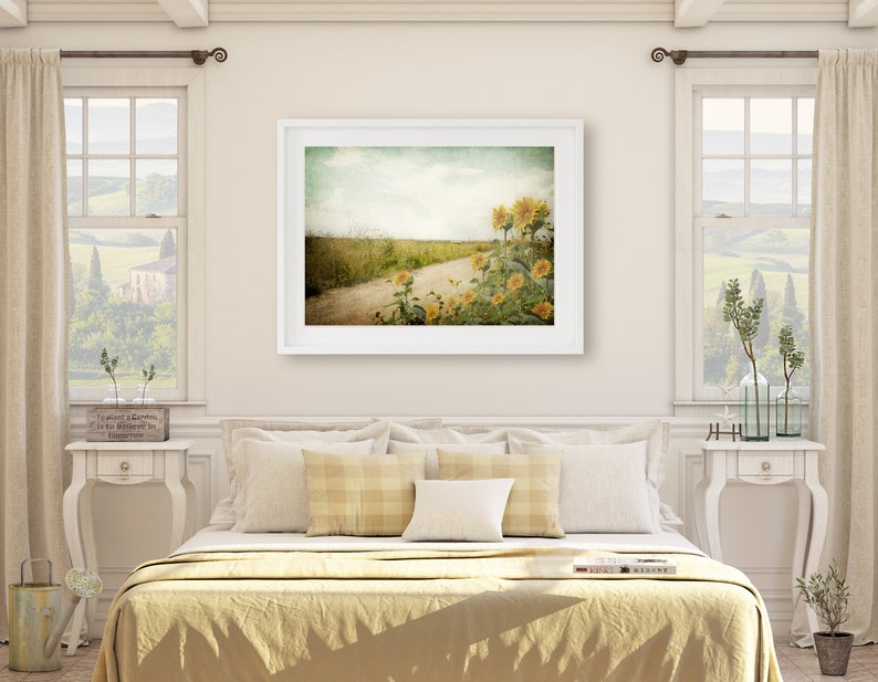 Puede incluir: Una impresi&oacute;n enmarcada en blanco de una fotograf&iacute;a de estilo vintage de un camino de tierra que conduce a trav&eacute;s de un campo de girasoles. El cielo es azul claro con nubes blancas. Los girasoles est&aacute;n en flor y el campo es verde.