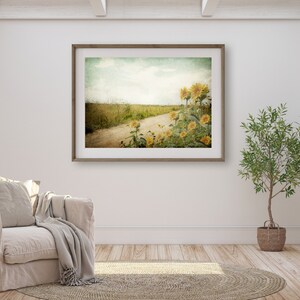Puede incluir: Una fotograf&iacute;a enmarcada de un camino de tierra que conduce a trav&eacute;s de un campo de girasoles. La foto tiene un aspecto vintage, descolorido y se muestra en una pared blanca en un ambiente de sala de estar.
