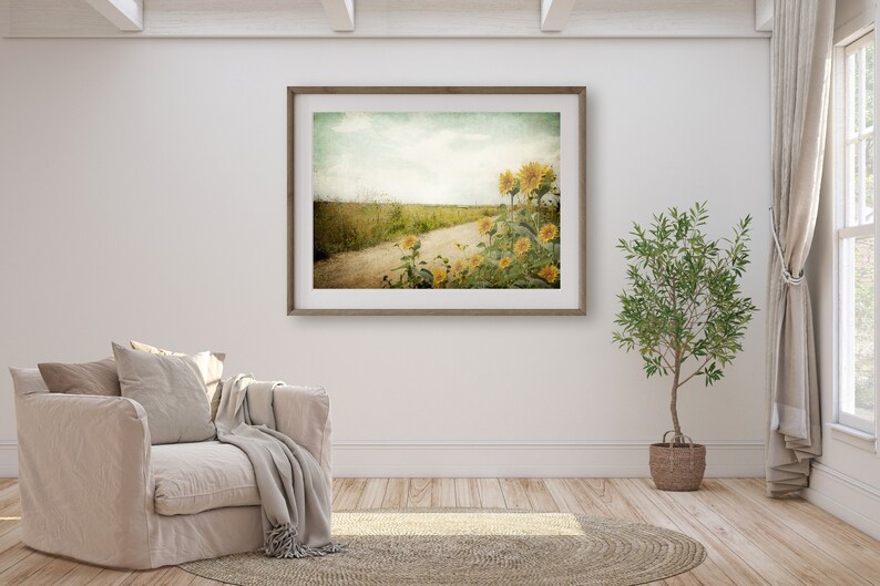 Puede incluir: Una fotograf&iacute;a enmarcada de un camino de tierra que conduce a trav&eacute;s de un campo de girasoles. La foto tiene un aspecto vintage, descolorido y se muestra en una pared blanca en un ambiente de sala de estar.