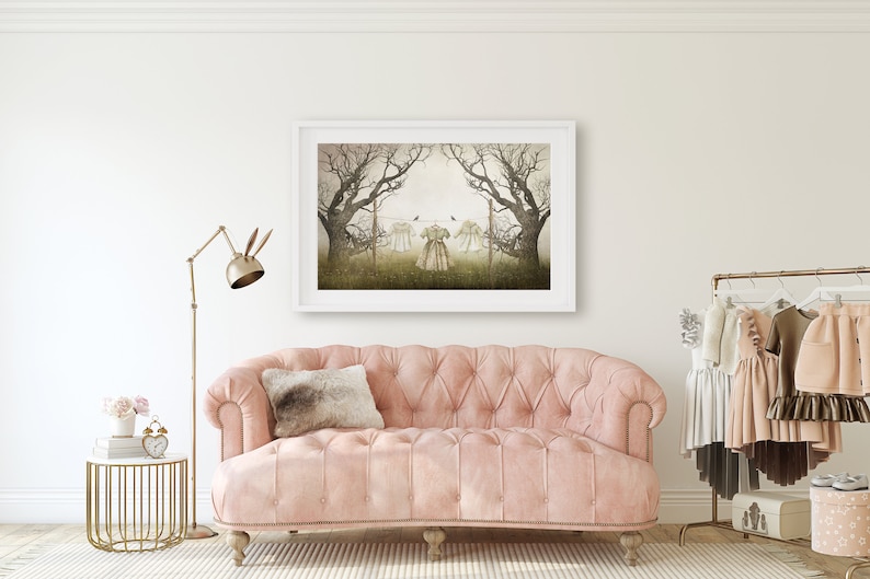 May include: A pink tufted sofa with a brown throw pillow in front of a white wall with a framed print of a foggy forest scene with three dresses hanging on a clothesline.