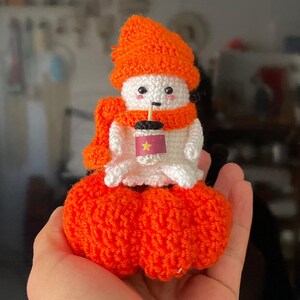 May include: A white crocheted amigurumi doll wearing an orange hat and scarf, holding a brown and white cup with a star on it. The doll is sitting on two orange crocheted pumpkins.