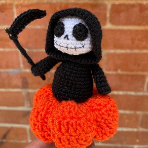 Op de afbeelding: Een handgemaakte gehaakte Halloween-decoratie met een Grim Reaper-figuur. De figuur heeft een zwarte capuchon, een wit schedelgezicht en een oranje pompoenvormige basis. Het houdt een kleine zwarte zeis vast.