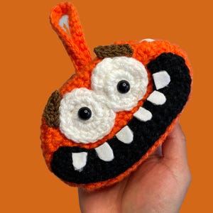 Puede incluir: Monstruo de crochet naranja con una gran sonrisa de dientes, ojos blancos con pupilas negras y cejas marrones.