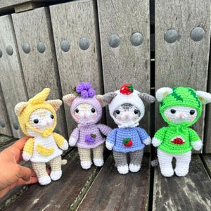 Puede incluir: Cuatro muñecos de gato de ganchillo hechos a mano, cada uno con un atuendo con temática de frutas. Los muñecos son blancos, grises y tostados, con atuendos en amarillo, morado, azul y verde. Los muñecos se exhiben sobre una superficie de madera.