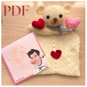 May include: A handmade crochet bear with a gray scarf and two crochet hearts, one red and one pink. The bear is on a cream-colored crochet blanket. A pink card with the words "Love You!" and a portrait of a person is on the left.