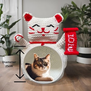 May include: A white and red crochet cat bed with a diameter of 9.8 inches. The cat bed is shaped like a Japanese Maneki Neko cat, with a red scroll that says "IOU". A cat is inside the bed.