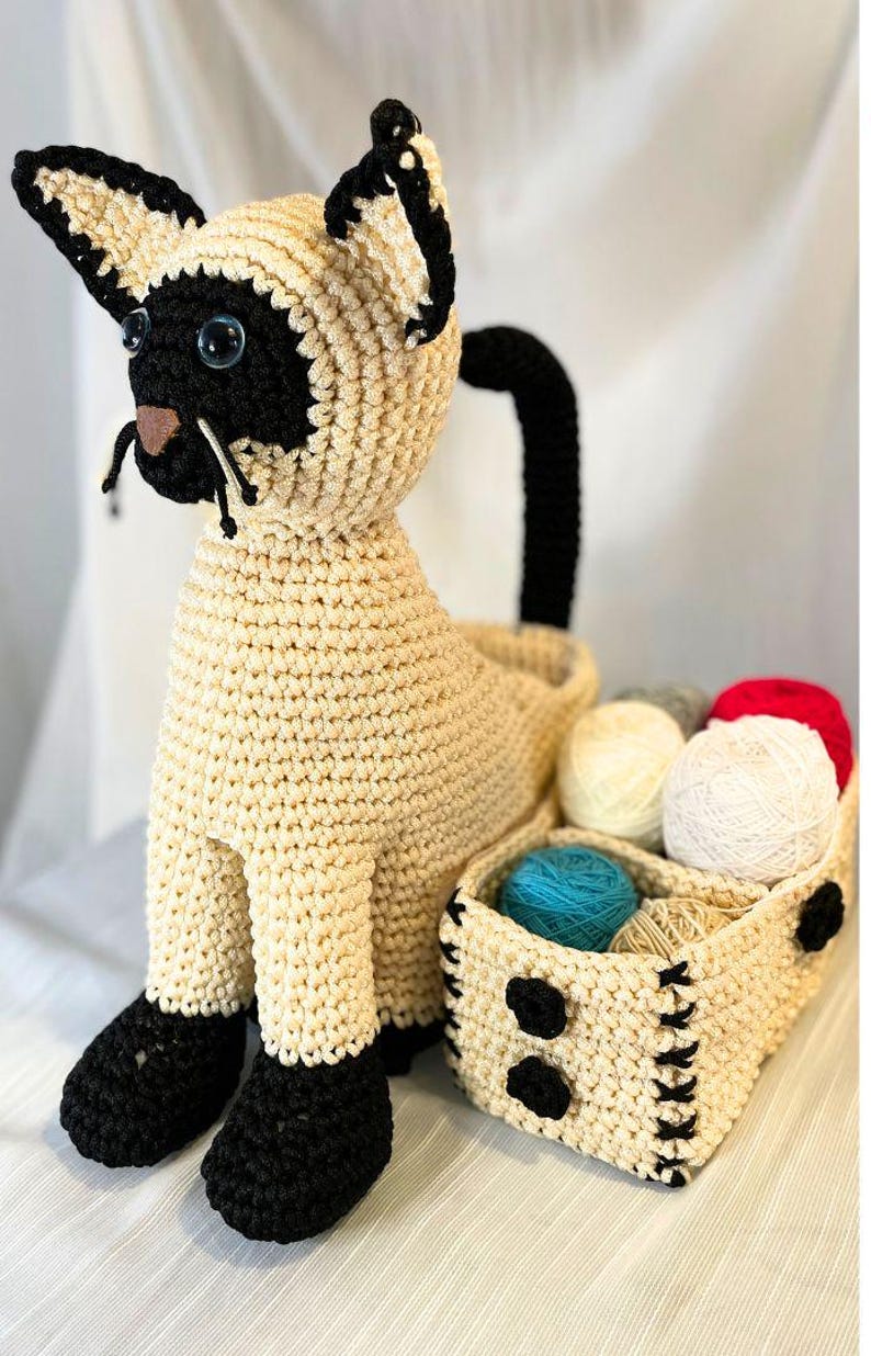 May include: A crocheted Siamese cat-shaped yarn basket. The cat has cream-colored fur with black accents on the face, ears, and paws. A black tail curves upwards. A small basket holds various yarn balls in colors like white, red, and blue.