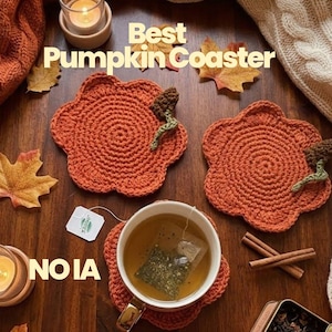 Puede incluir: Tres posavasos de ganchillo con forma de calabaza naranja, con tallos marrones y zarcillos verdes. El texto "Best Pumpkin Coaster" y "NOIA" es visible. Los posavasos están dispuestos con una taza de té, una bolsita de té y hojas.
